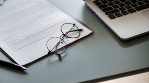 Document with reading glasses resting on it, sitting next to a laptop. Keyword: Business plan